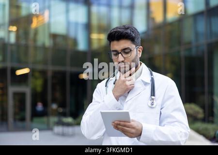 Young male doctor in a lab coat and stethoscope seriously looking at a digital tablet, deep in thought, representing modern medical technology and professional problem-solving outdoors Stock Photo