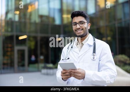Young indian male doctor in a white lab coat and stethoscope holding a digital tablet and smiling confidently ,while standing outside a modern clinic or hospital building Stock Photo