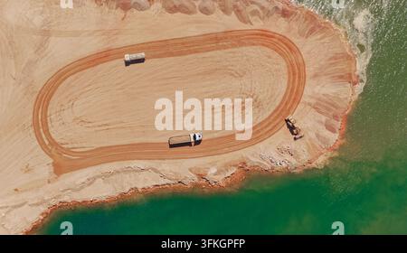 Elevated drone view capturing an active construction site with dump trucks, excavators, and heavy machinery working on sand extraction near a vibrant Stock Photo