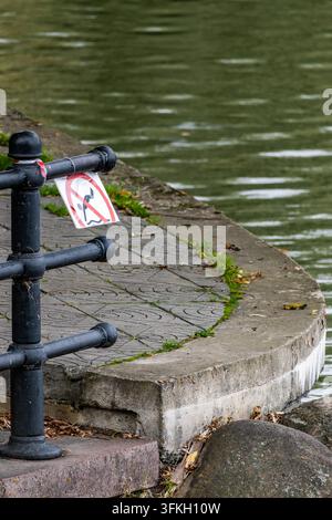 A black iron safety railing displaying a red pictogram sign prohibiting swimming stands beside a concrete pathway at the water edge. The paved walkway Stock Photo