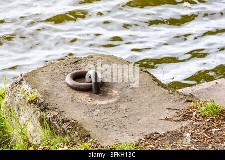 A large rusted iron mooring ring is secured to a weathered concrete block on a waterfront bank. Calm water ripples in the background behind the concre Stock Photo