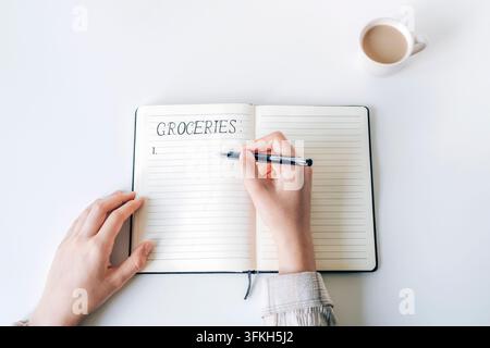 Person writing a grocery list in an open notebook beside a cup of coffee. Top view, flat lay. Stock Photo