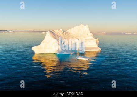 Aerial view of a large iceberg floating in the deep blue ocean, illuminated by the warm golden light of the setting sun with reflections on the water Stock Photo