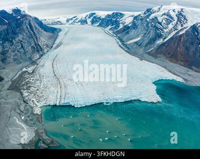 Aerial view of a massive glacier flowing into a turquoise glacial lake, surrounded by rugged snow-capped mountains and rocky terrain in Greenland. Stock Photo