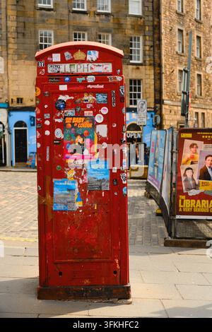 Edinburgh, Scotland - August 2 2026 - Red sticker covered telephone booth stands on sunlit cobblestones with old stone buildings behind. Stock Photo
