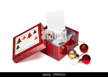 Red gift box opens to reveal wrapped item and blank card. Bright red and gold accents frame festive holiday theme. Three shiny baubles lie beside box, Stock Photo
