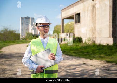 Architect in hard hat and safety vest holding closed laptop at an unfinished building site. Architectural services construction management and office- Stock Photo
