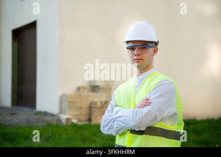Construction supervisor wearing safety gear stands with crossed arms beside a building renovation site. Architectural services construction leadership Stock Photo