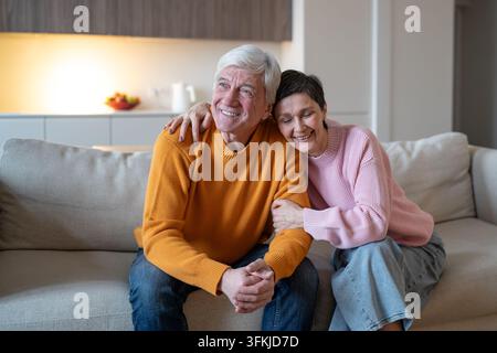 Cheerful senior wife hugs laughing husband as elderly couple sits together on couch at home Stock Photo