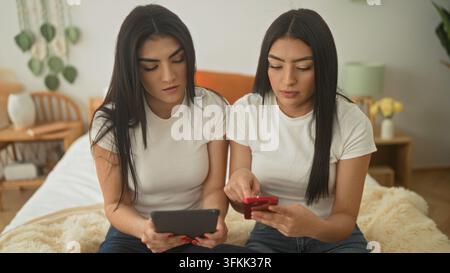 Two women sisters sit on bed, one points finger to tablet while the other holds a red smartphone and scrolls in a bedroom in a building; calm focused. Stock Photo