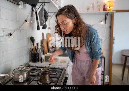 Interested woman preparing Turkish black coffee in cezve on gas stove during morning routine at home Stock Photo