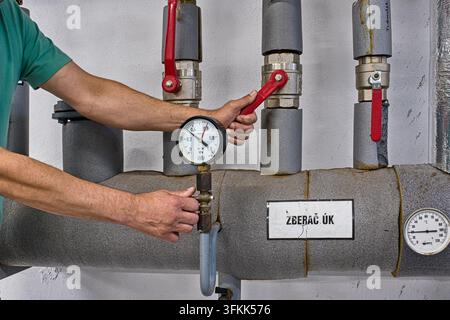 Technician operating a control valve and pressure gauge on insulated central heating collector pipes in a boiler room, heating service. Stock Photo