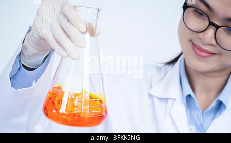 Erlenmeyer flask filled with orange liquid chemical held and examined by young female researcher scientist in laboratory Stock Photo