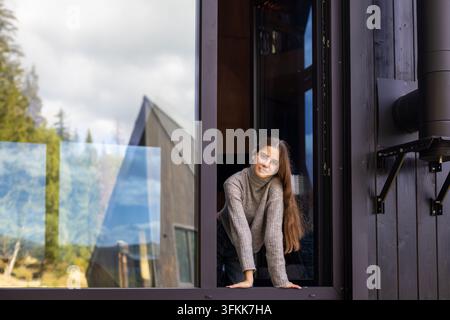 Young Woman Leaning Through Modern Cabin Window. Young woman leans through an open window of a modern wooden cabin, looking at the camera with forest Stock Photo
