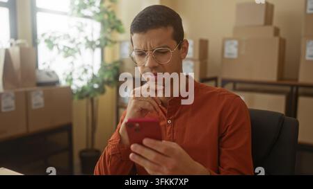 Man holding smartphone, hand on chin, studying a message amid stacked shipping boxes and labels in a building; concern order logistics. Stock Photo