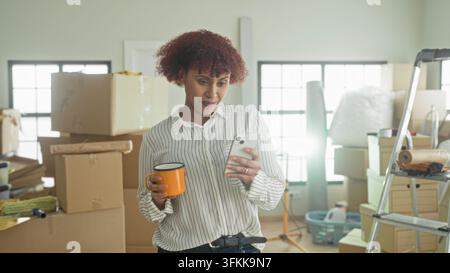 Woman holds smartphone and orange mug, hand tapping phone while surrounded by moving boxes in building; settling in contentment. Stock Photo