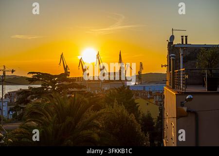 Shipyard Cranes at Sunset Over Coastal City. Golden sunset illuminates shipyard cranes above a coastal city, with rooftops, trees and a terrace in the Stock Photo