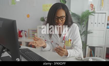 Woman doctor in labcoat tapping smartphone screen with thumb while seated at desk with computer and stethoscope visible in clinic building; focused em Stock Photo