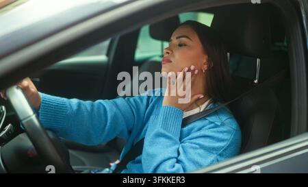 Woman wearing blue sweater touching cheek and steering car on street, seatbelt fastened; discomfort concern fatigue. Stock Photo