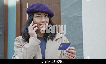 Woman on phone holding creditcard at building entrance near intercom and door, wearing purple beret and beige coat while checking card; payment verifi Stock Photo