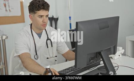 Man doctor with stethoscope leans on arm and uses computer mouse at clinic desk amid paperwork, medicine bottle and anatomical poster; concentration. Stock Photo