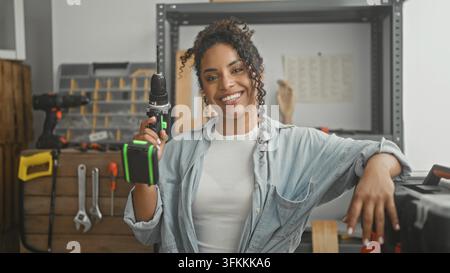 Woman holding a cordless drill upright, right hand gripping the drill and left hand resting on toolbox inside a workshop building; confidence craft sk Stock Photo