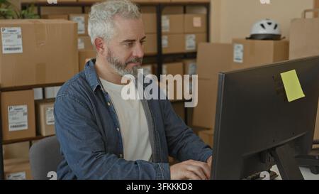 Man typing on computer with packages and stacked boxes, hands on keyboard in a packing area inside a building; focused efficiency. Stock Photo