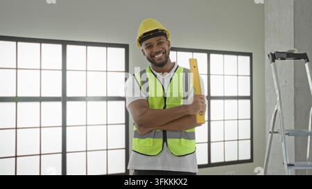 Man builder in hardhat smiles and holds a yellow measuring level with arms crossed revealing forearm inside a building; confidence professionalism. Stock Photo