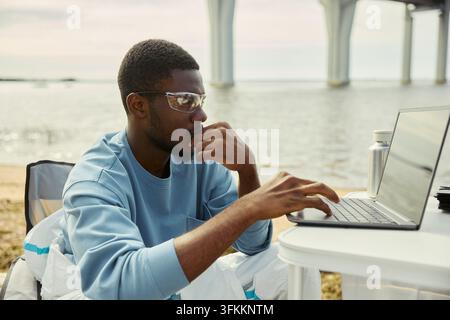 Focused Black Man Working on Laptop Outdoors by River Stock Photo
