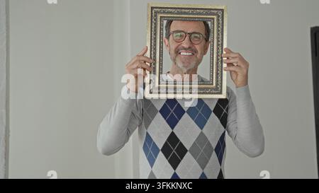 Man holds ornate picture frame over face, smiling and wearing an argyle sweater in a building interior hall; self reflection joy. Stock Photo