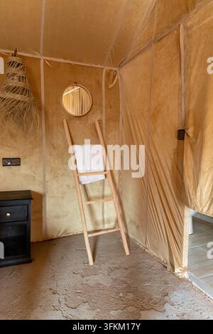Rustic beige canvas tent interior with a wooden ladder towel rack, round mirror, woven lamp shade and dark bedside cabinet. Stock Photo