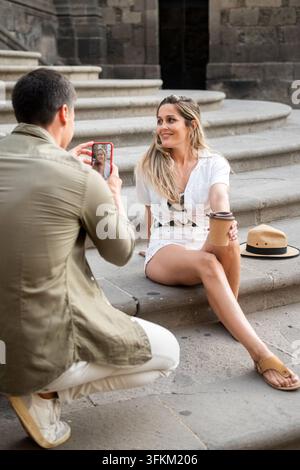 Male tourist using a smartphone to photograph his partner sitting on stone steps during a city sightseeing trip. Stock Photo