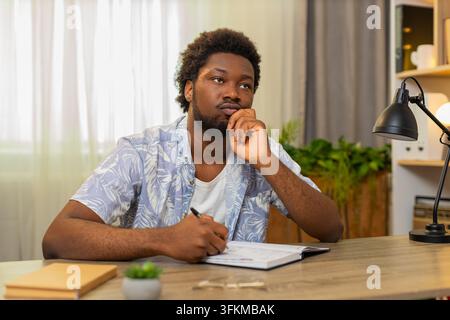 African American young man on home table writes plans in notebook and lists tasks for daily work. Black guy on home table sets goals and creates schedule with focused mood for better time management. Stock Photo