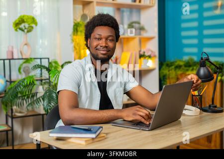 African American young man on home table opens laptop for remote work then starts typing with focused calm mood. Black guy on home table uses computer for freelance task online web job search indoors. Stock Photo