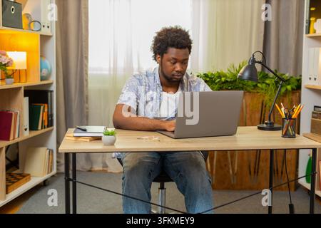 African American young man on home table checks email on laptop and reads important letter with focused face. Black guy on home table thinks about message and starts typing a careful reply from home. Stock Photo