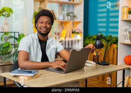 African American young man on home table opens laptop for remote work then starts typing with focused calm mood. Black guy on home table uses computer for freelance task online web job search indoors. Stock Photo