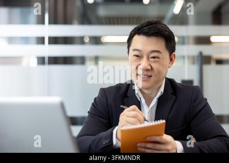 Businessman taking notes during a video conference, professional man using a laptop for financial analysis and corporate communication. Executive working at a desk in a modern office setting. Stock Photo