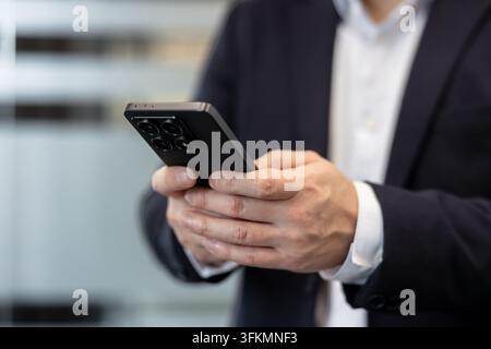 Businessman using a smartphone for digital communication and corporate networking tasks. Professional man in a suit holding a mobile phone to manage business operations in a modern office environment. Stock Photo