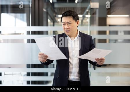 Financial analysis and business stress, professional man reading documents with concern in office. Corporate worker reviewing paperwork with deadline pressure and anxiety during a busy workday. Stock Photo