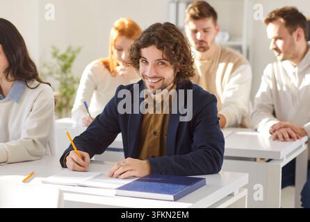 Cheerful curly haired male student writing down notes while having lesson in class Stock Photo