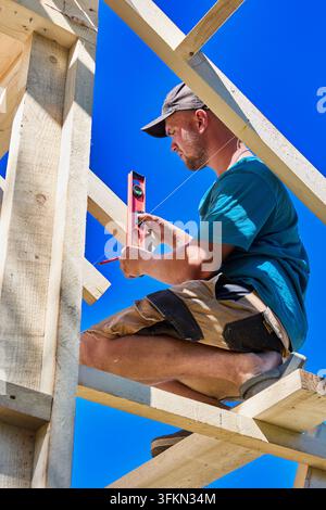 Wooden makeshift scaffolding holds carpenter as he aligns wooden roof truss using pocket level during framing. Stock Photo