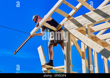 Spirit level guides young construction worker aligning roof rafter while standing on wooden makeshift scaffolding. Stock Photo