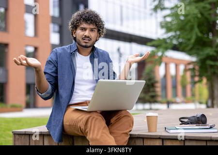 Confused student with laptop on university campus, young man having trouble with online education assignment or exam preparation while sitting outdoors at a college library area. Stock Photo