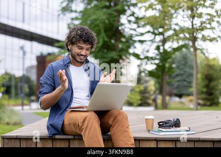 Happy student attending an online university lecture on a laptop at a campus park. Young man studying with books and coffee during remote education course preparation for an upcoming academic exam. Stock Photo