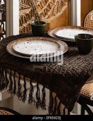 Rustic table setting with stacked speckled ceramic plates, dark green cups, and a fringed dark woven runner. Stock Photo