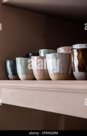 Row of colorful handcrafted ceramic cups with unglazed bases arranged on a light wooden shelf. Stock Photo