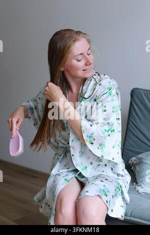 Woman grimacing while detangling wet hair at home, a concept of bad hair days, breakage prevention and beauty frustration. Stock Photo