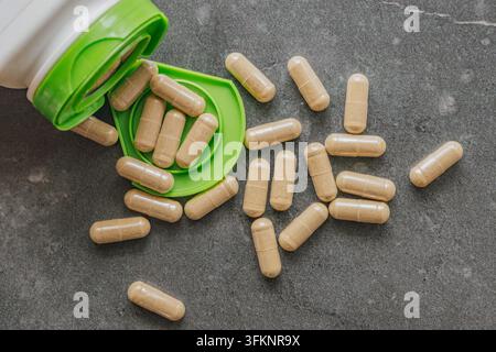 Herbal vitamin capsules spilling out from open white plastic jar on dark grey slate surface for medical healthcare concept. Stock Photo