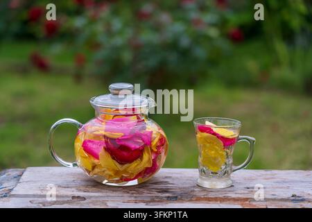 Beautiful and fragrant tea from yellow, pink and red petals roses in glass teapot and mug in the summer garden on wooden table. Close up herbal flower Stock Photo