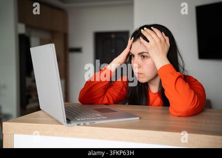 Frustrated woman holding her head while using laptop at desk in home office. Workplace stress technology frustration and deadline anxiety. Stock Photo